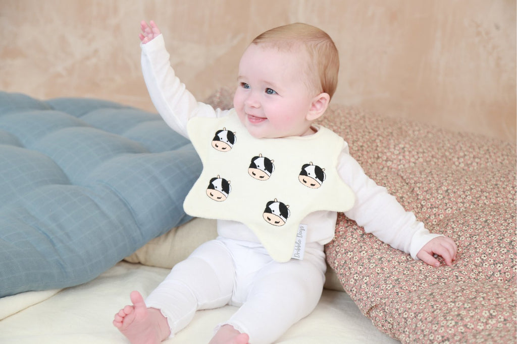 Baby wearing a cream, star shaped Dribble Days bib with embroidered Cows